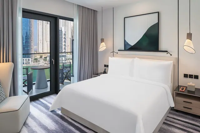 A bedroom with a made king bed, a white armchair, pendant lights and a glass door opening onto a balcony with a view of high-rise towers.