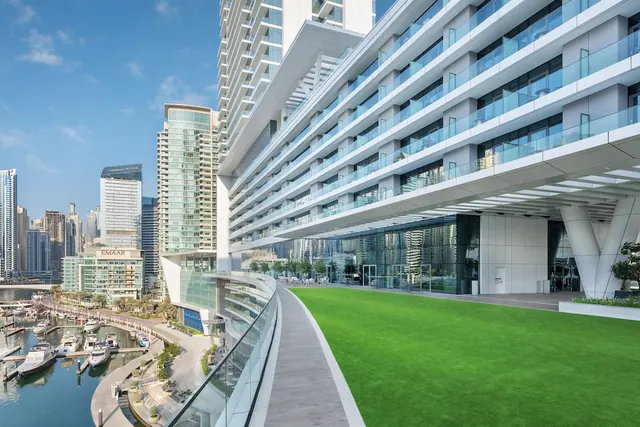 The exterior of a white and glass tower with glass balcony railings and a green lawn, with a marina and high-rise skyline behind it.