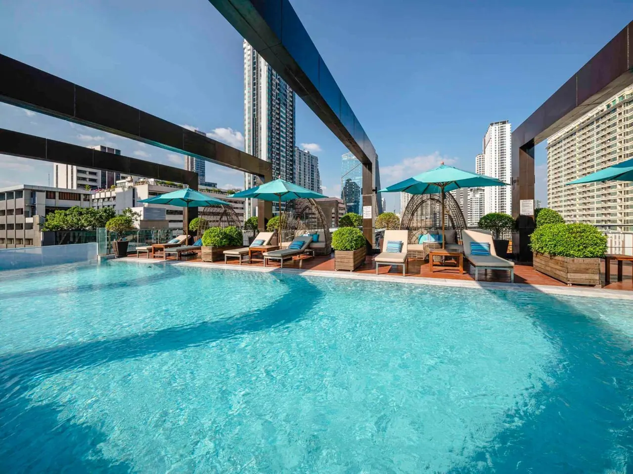 A rooftop swimming pool with turquoise water, teal umbrellas, sun loungers and potted shrubs, with high-rise buildings visible beyond.