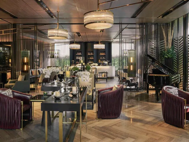 A hotel lounge with pendant chandeliers, burgundy leather armchairs, a grand piano, and a bar counter with bottle shelves in the background.