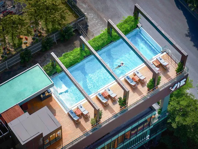 Aerial view of a rooftop swimming pool with one swimmer, sun loungers on a wooden deck, and a glass railing above a city street with trees.