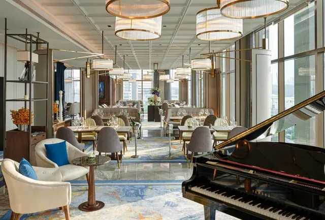 A dining room with tables set for service, upholstered armchairs and banquettes, a black grand piano in the foreground, and floor-to-ceiling windows overlooking city buildings.