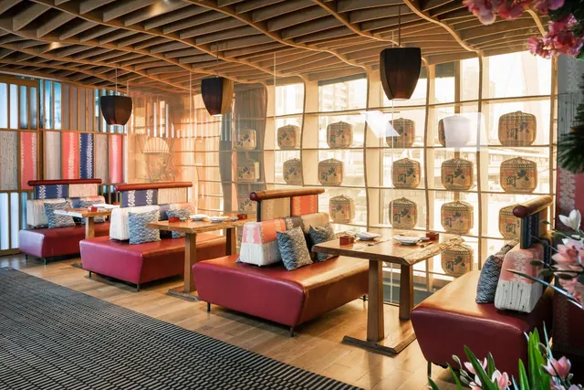 A restaurant dining area with red leather booth seating, wooden tables, pendant lights and a large window showing blurred city buildings outside.