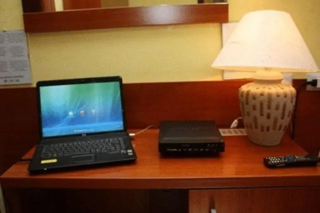 A wooden desk holds a open laptop, a black set-top box, a remote control and a cream ceramic table lamp against a yellow wall.