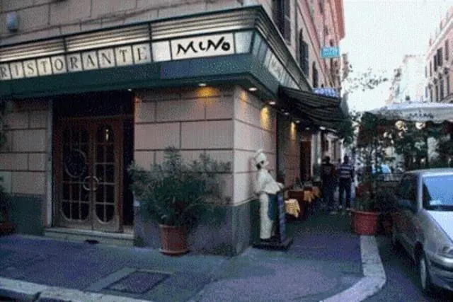 Exterior of Ristorante Mino on a street corner, with a chef figure statue beside the entrance, potted plants, a parked silver car, and two people standing in the background.