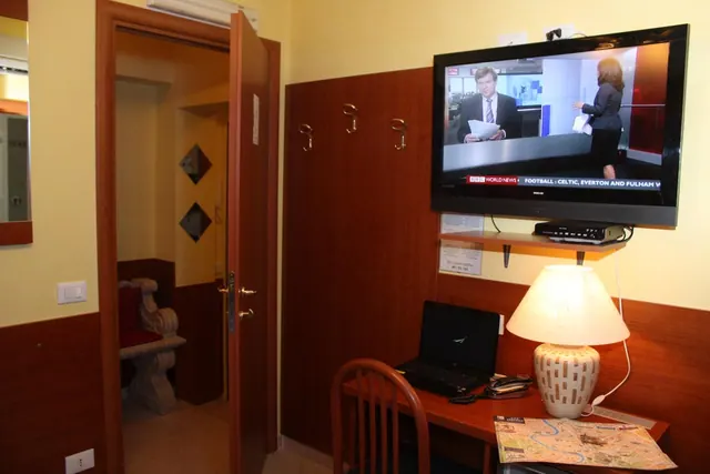 A hotel room with a wall-mounted television showing a news broadcast, a desk holding a laptop, a lamp and a folded map, and an open door onto a corridor.