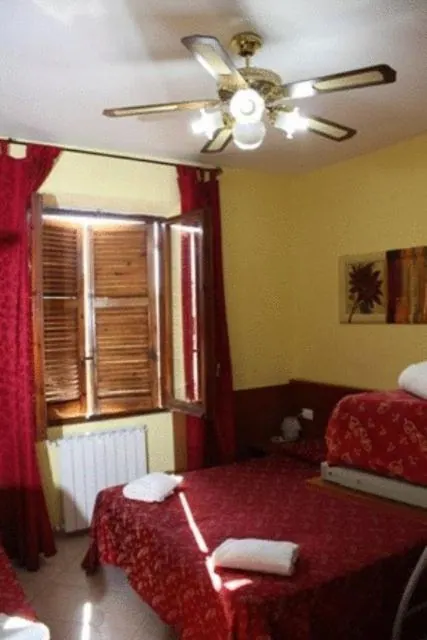 A bedroom with two beds covered in red floral spreads, red curtains, open wooden shutters and a ceiling fan.