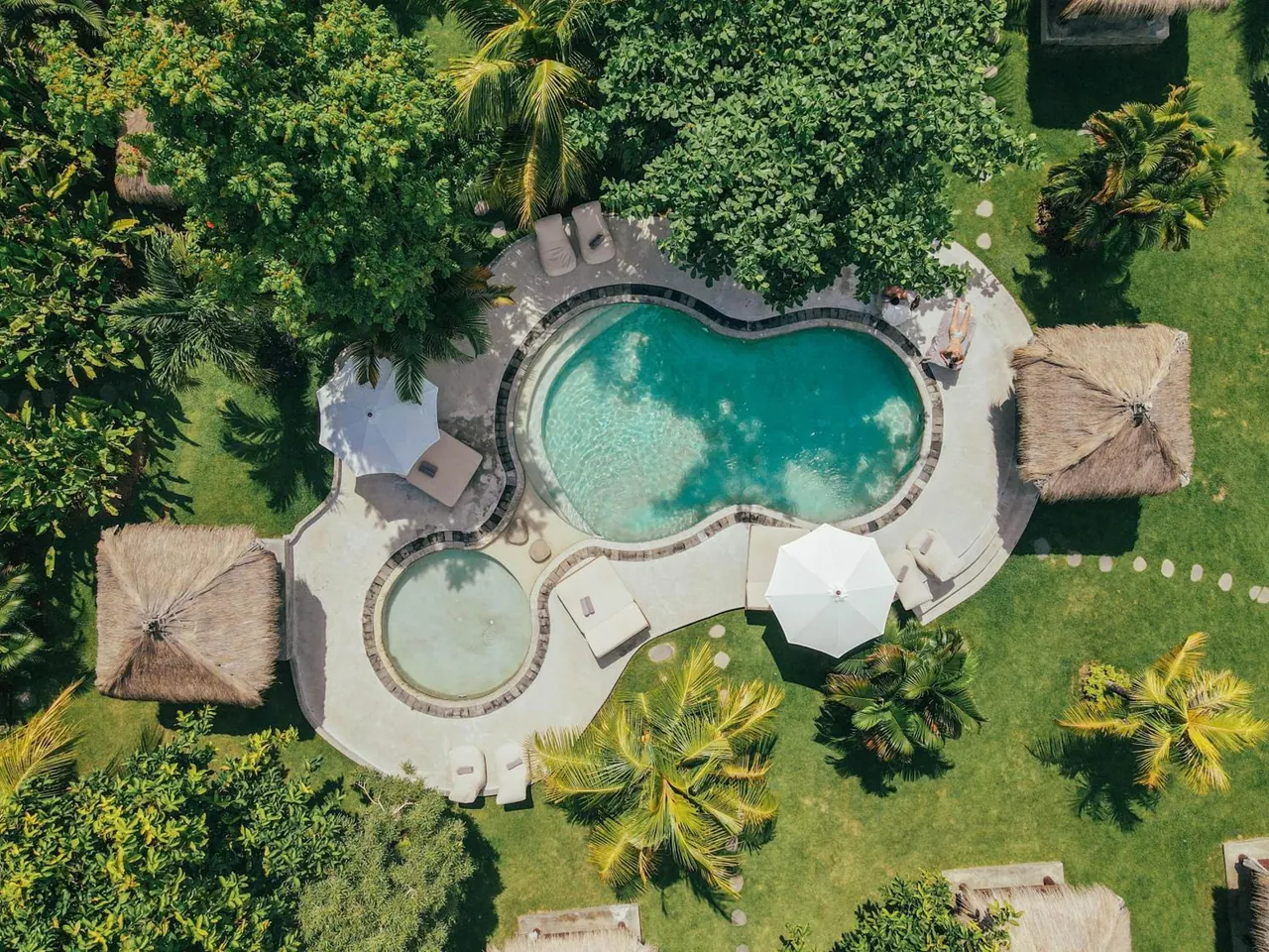 Aerial view of a kidney-shaped pool with a round plunge pool, sun loungers, white umbrellas and thatched gazebos among palm trees on a lawn.