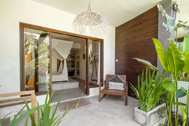 A covered terrace outside a bedroom has glass sliding doors open onto a bed with white mosquito netting, a wooden chair with a patterned cushion, and potted plants on a concrete floor.