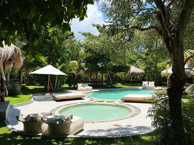 A swimming pool and a small round pool sit on a paved deck surrounded by garden, with sun loungers, beanbag seats and a white parasol.
