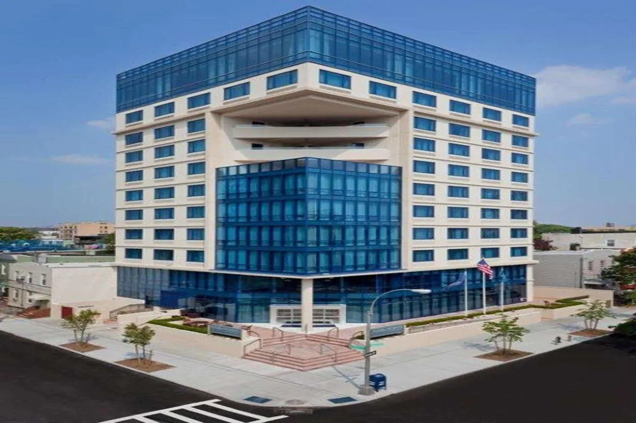 Exterior of a multi-storey hotel building with blue glass upper and lower sections, a stepped entrance and flagpoles on a street under a clear sky.