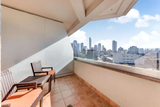 A tiled balcony with wicker chairs, orange cushions and a small table overlooks a city skyline under a blue sky.