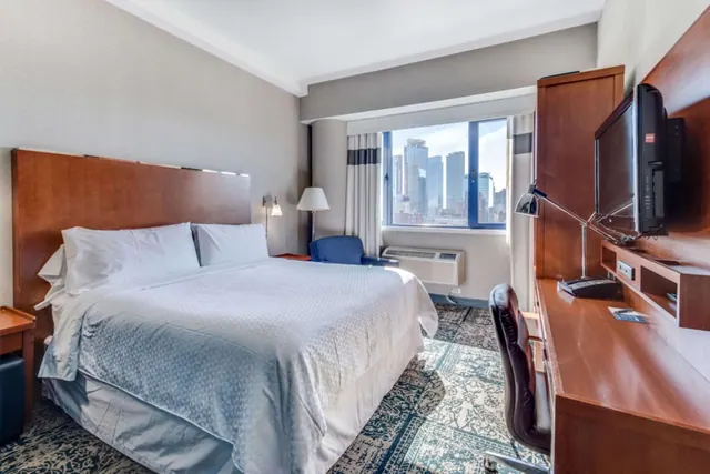 A hotel bedroom with a king bed, a wall-mounted television and a wooden desk, with a window showing a city skyline.
