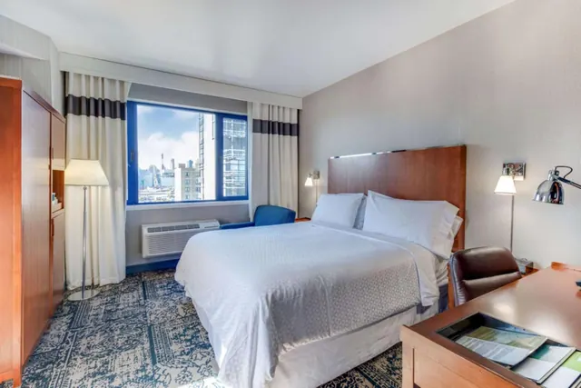 A hotel bedroom with a large bed, a wooden desk and leather chair, a blue armchair, and a window looking out onto city buildings.