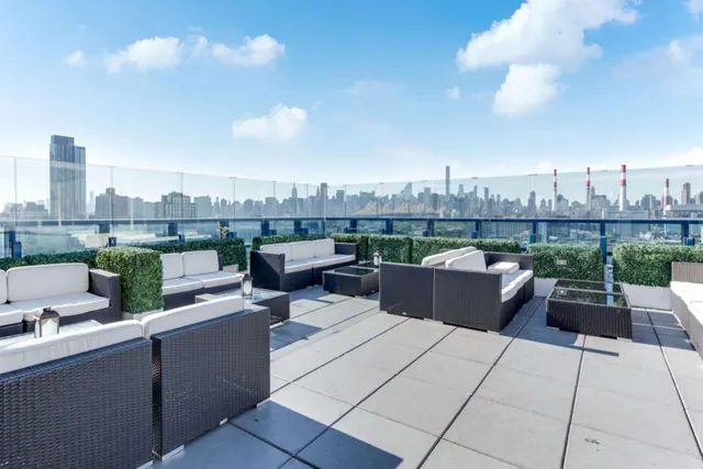 A rooftop terrace with dark wicker sofas and armchairs with white cushions, a glass railing, and a city skyline in the distance.