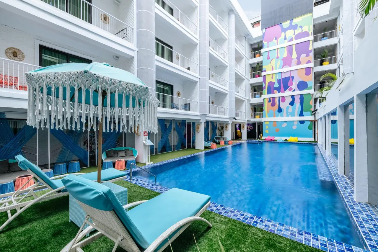 A long blue swimming pool in an open courtyard, with turquoise sun loungers and a fringed umbrella on the grass in the foreground and a colourful mural on the building beside it.