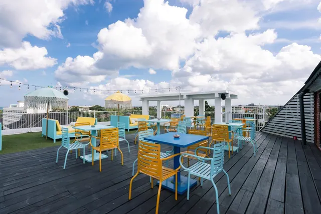 A rooftop terrace with blue and yellow tables and chairs, lounge seating under fringed umbrellas, and a white pergola, with low buildings visible beyond the railing under a cloudy sky.