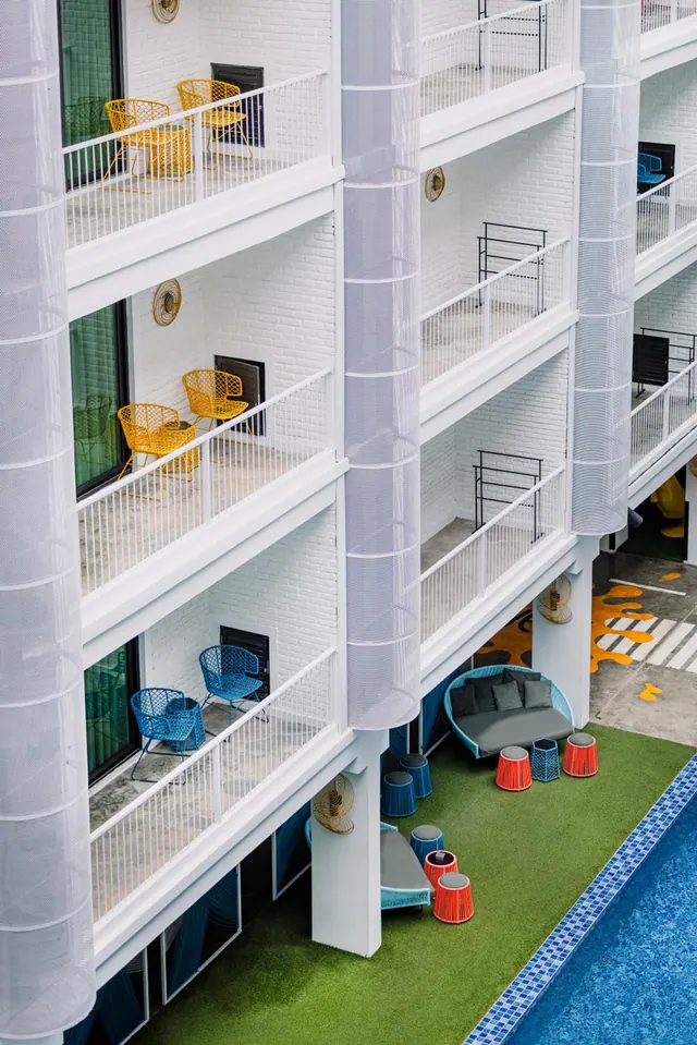 Upper-floor balconies with yellow and blue chairs overlook a courtyard with green turf, a blue daybed, colourful stools and the edge of a pool.