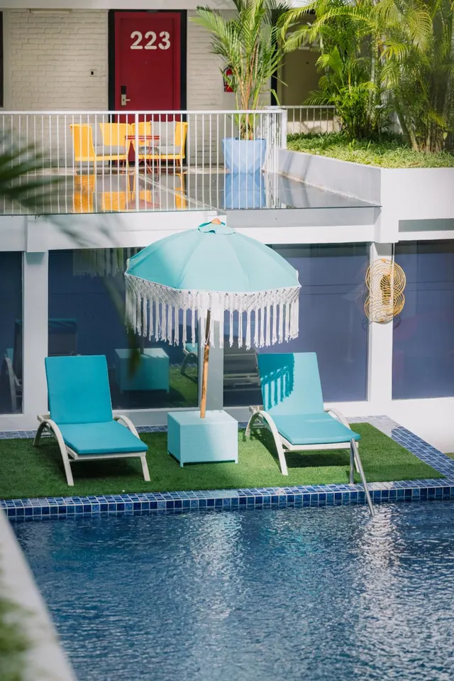 Two turquoise sun loungers and a fringed turquoise umbrella stand on artificial grass beside a blue-tiled pool, with a balcony and a red door numbered 223 above.
