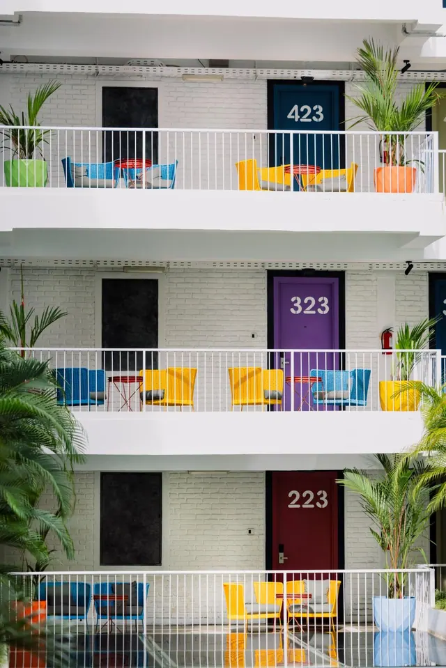 A white multi-storey building facade shows balconies furnished with coloured chairs and potted palms, with brightly painted doors numbered 423, 323 and 223.