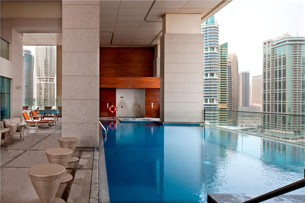 A pool with a glass edge overlooks high-rise towers, with sun loungers and wicker stools on the covered deck to the left.