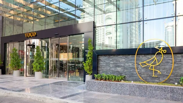 The glass-fronted entrance of a VOCO hotel with potted topiary plants, a sliding door and a gold bird logo on a stone wall.