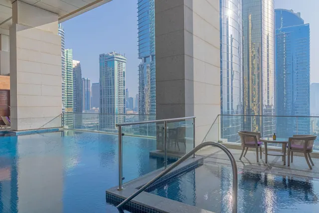 A pool with a glass railing and a small plunge pool sit beside a terrace with two chairs and a table, overlooking high-rise towers.