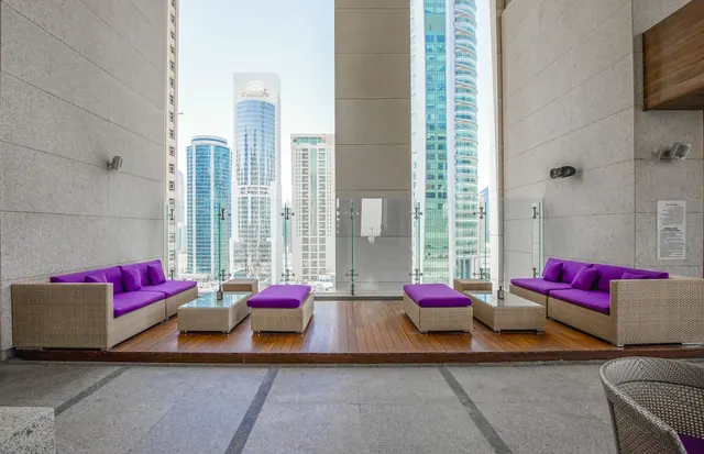 Wicker sofas with purple cushions and low glass-topped tables sit on a wooden deck, with high-rise towers visible through the glass barrier behind them.