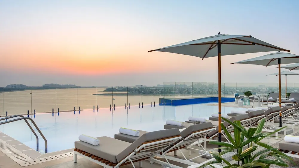 An infinity pool with grey sun loungers and white umbrellas overlooks the sea and a distant shoreline at sunset.