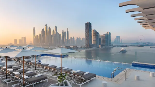 A rooftop infinity pool with sun loungers and white parasols looks out over a city skyline, a waterway and the Ain Dubai observation wheel.