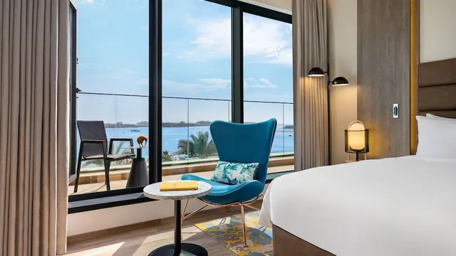 A bedroom with a made king bed, a blue armchair and floor-to-ceiling windows opening onto a balcony with a view of the sea.