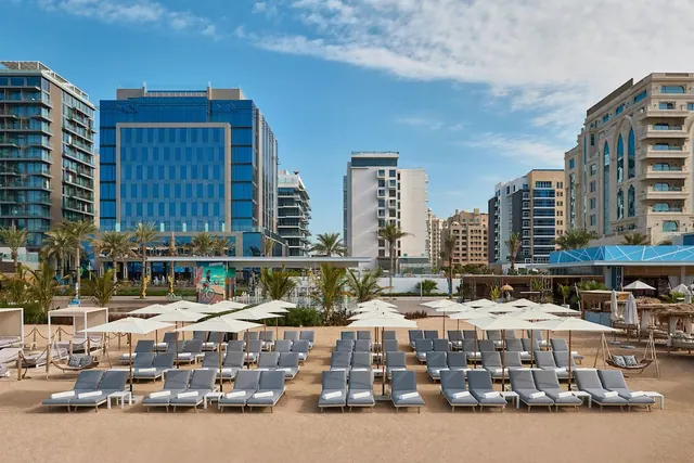 Rows of grey sun loungers and white parasols stand on a sandy beach, with high-rise hotel and apartment buildings behind them.