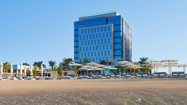 A blue glass hotel tower stands behind a sandy beach with rows of sun loungers and white umbrellas.