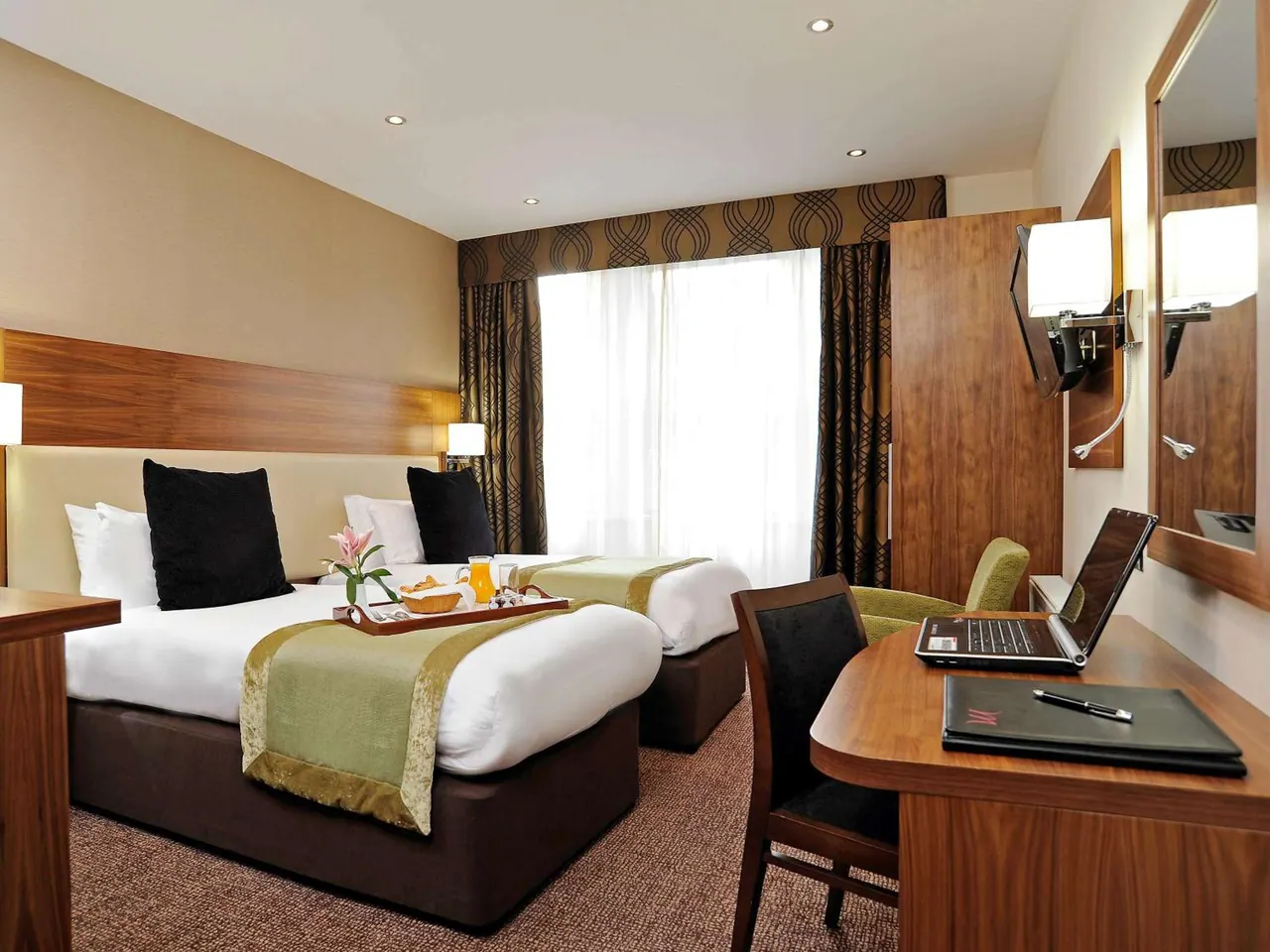 A twin-bedded hotel room with a breakfast tray on one bed, a wooden desk with a laptop and chair, a wooden wardrobe, and a wall-mounted television.