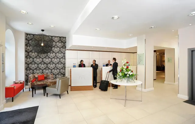 A hotel lobby with a reception desk where staff stand, a red bench and patterned sofa, and a floral arrangement on a round table.