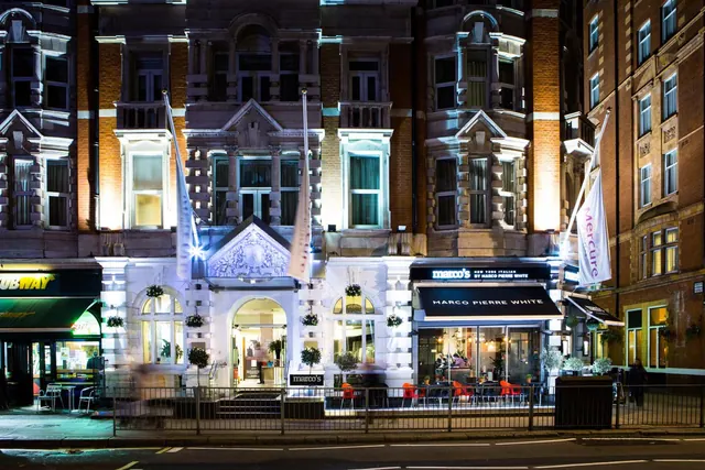 Night view of the building front with Marco's and Marco Pierre White signage, outdoor tables behind a railing, and Mercure flags.