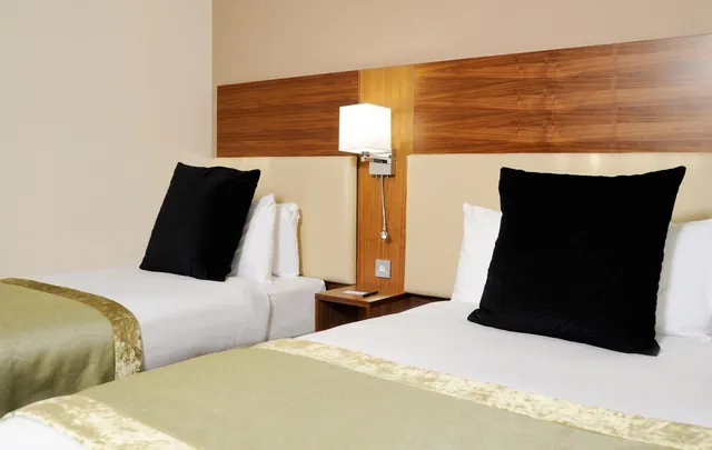 Two made-up beds with black cushions, a wall-mounted reading lamp and a wood-panelled headboard in a hotel bedroom.