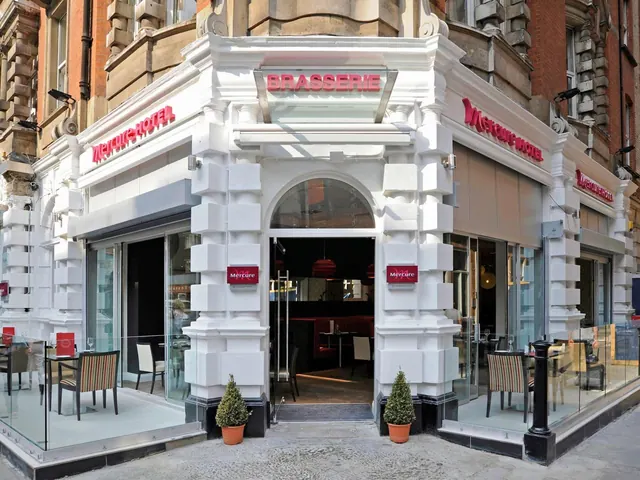The white corner entrance of a Mercure hotel with a Brasserie sign, glazed front seating with tables and chairs, and two potted topiary trees on the pavement.