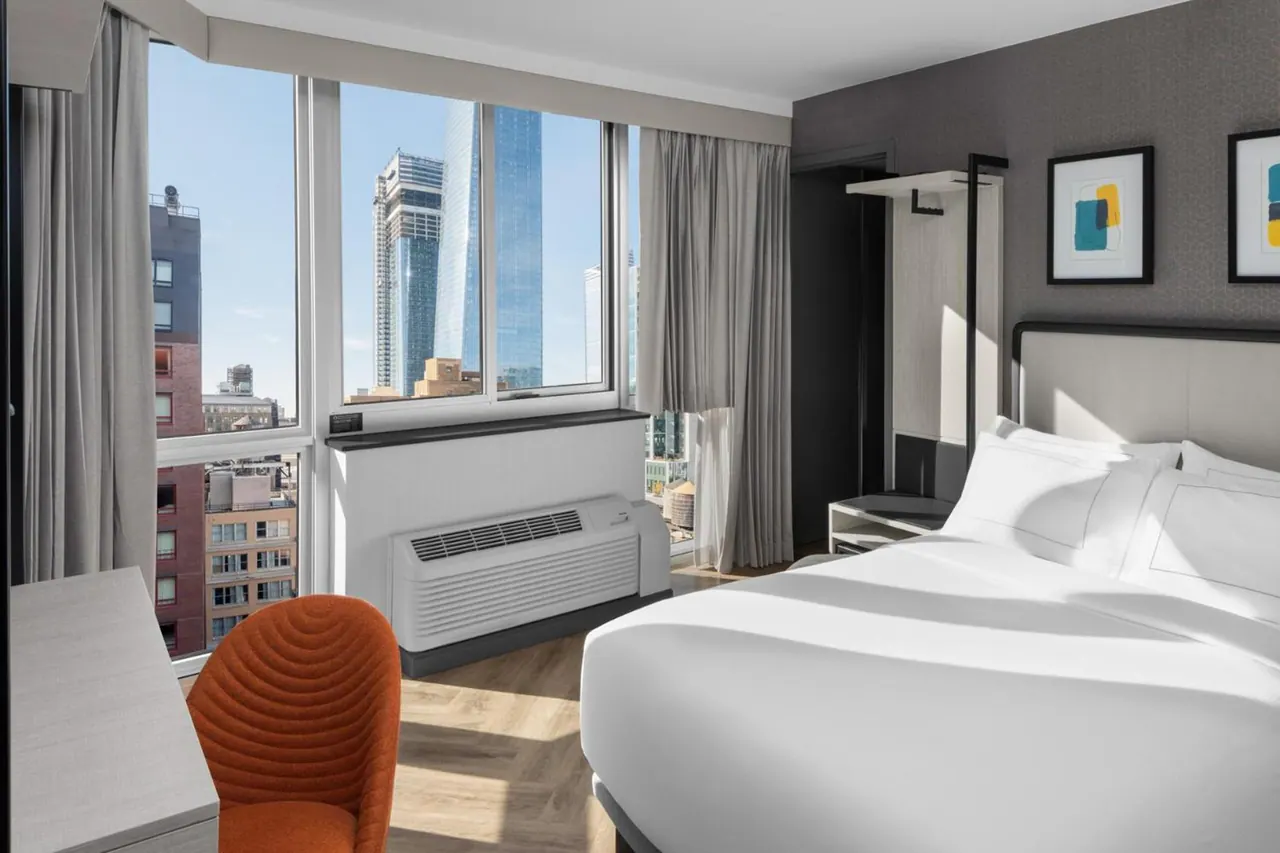 A hotel bedroom with a made king bed, a corner window looking out on city buildings, a wall-mounted air conditioning unit, and a desk with an orange chair.