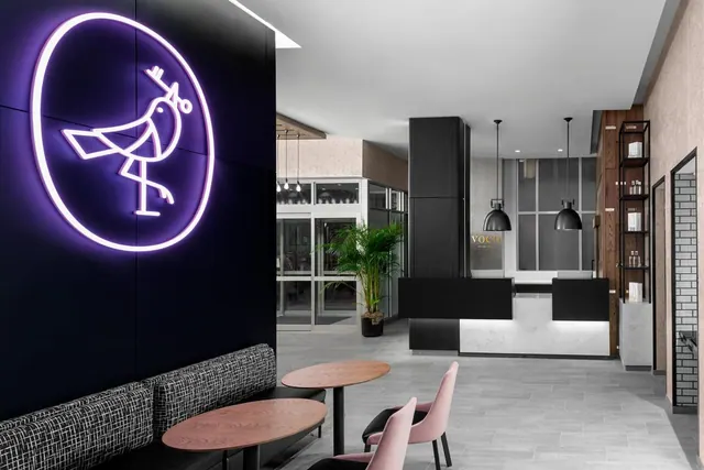 A hotel lobby with a glowing purple neon bird logo on a black wall, a banquette seating area with round wooden tables and pink chairs, and a reception desk in the background.