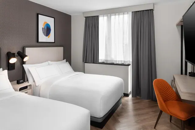 A hotel bedroom with two beds dressed in white linen, wall-mounted reading lamps, a window with grey curtains, an orange chair at a desk, and a wall-mounted television.