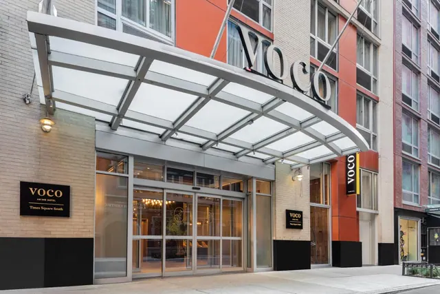The entrance of the VOCO Times Square South hotel, with a curved glass canopy above glass doors and the hotel name on the facade.