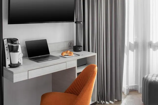 A hotel room corner with a wall-mounted television above a desk holding a laptop, a plate of croissants and a coffee maker, beside an orange chair and grey curtains.