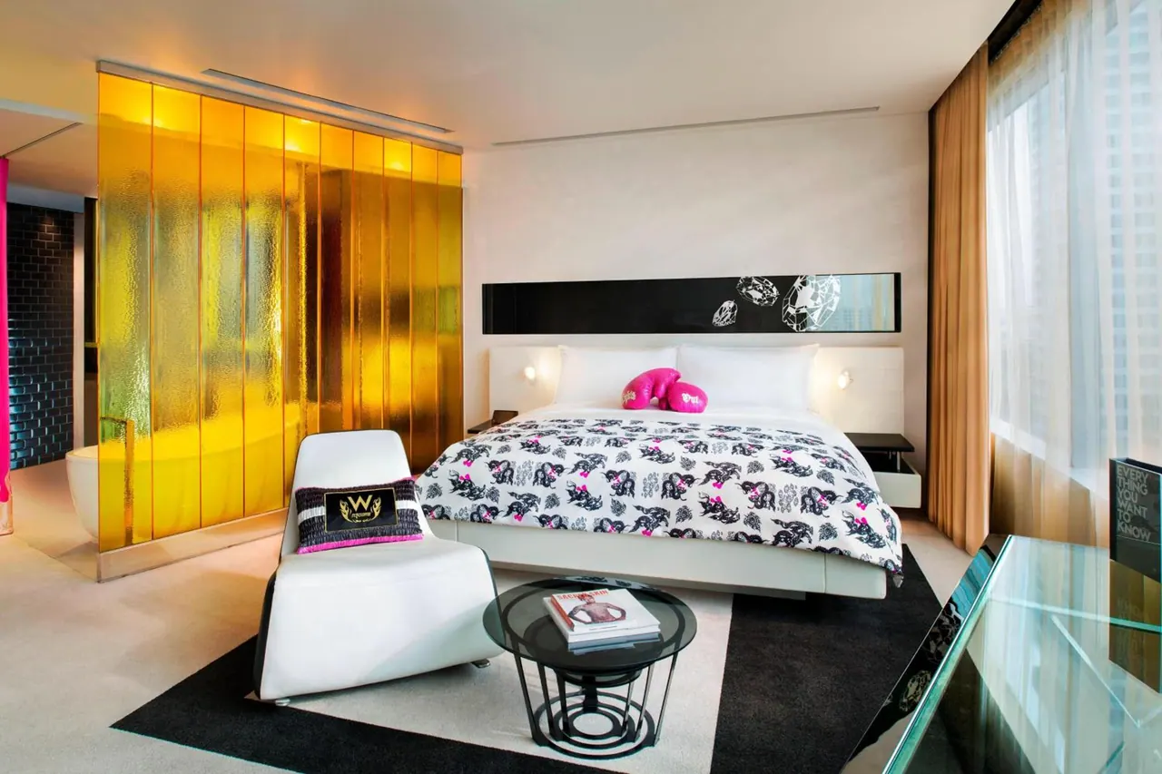A king bed with a black and white patterned duvet and pink cushions sits beside an amber glass partition, with a white lounge chair and round black coffee table in the foreground.