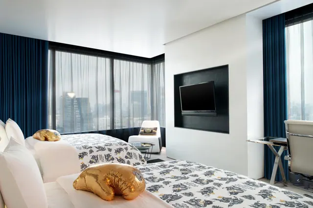 A hotel bedroom with two beds in patterned white bedding, a wall-mounted television, a white armchair and a desk, with floor-to-ceiling windows showing a city skyline.