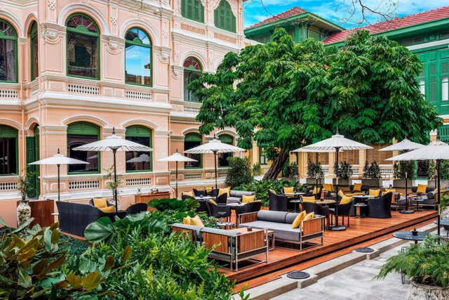 An outdoor lounge terrace with grey sofas, yellow cushions, white parasols and dining tables sits in front of a pink colonial-style building with green shutters.