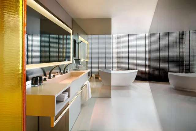 A bathroom with a long trough basin and deck-mounted faucets beside two freestanding bathtubs, with horizontal blinds along the back wall.