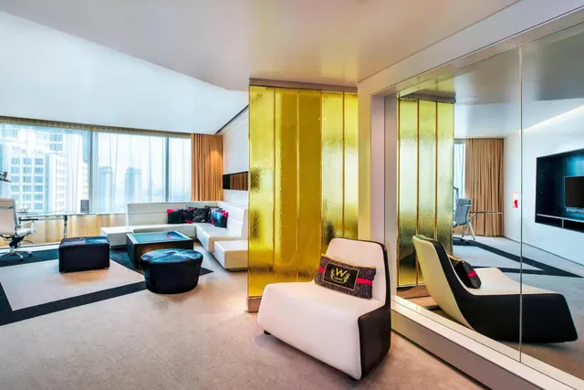 A suite living area with a white sectional sofa, black ottomans, a gold textured glass divider, a mirrored wall, and a window showing high-rise buildings.
