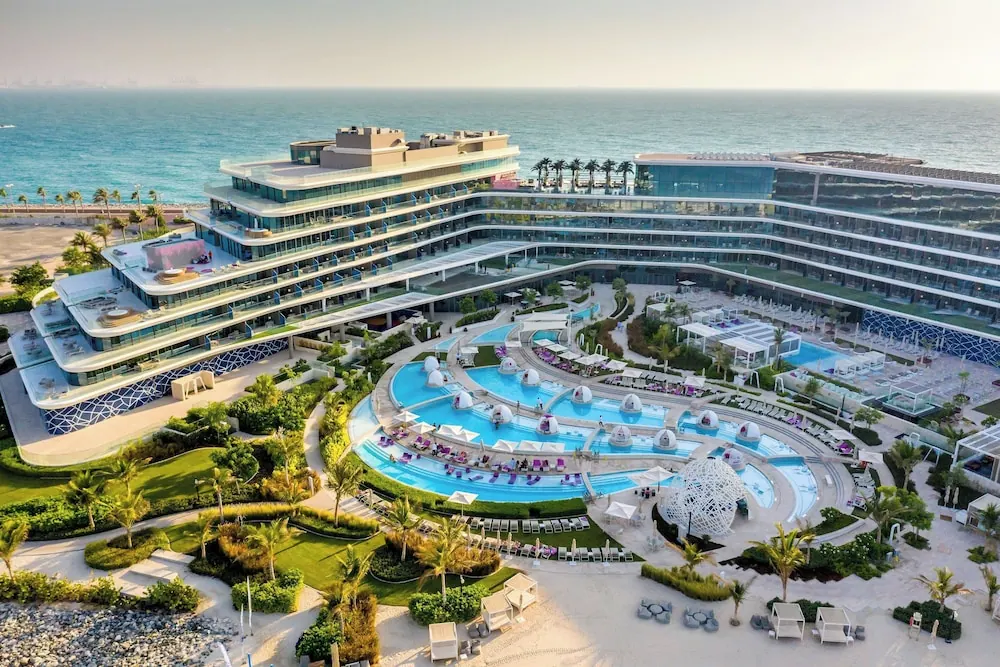 Aerial view of a resort with a multi-pool complex, sun loungers and umbrellas, with the sea and modern hotel buildings beside it.
