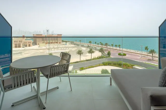 A furnished balcony with a round table, chairs and a daybed looks out over a coastal road, palm trees and the sea toward a hazy skyline.
